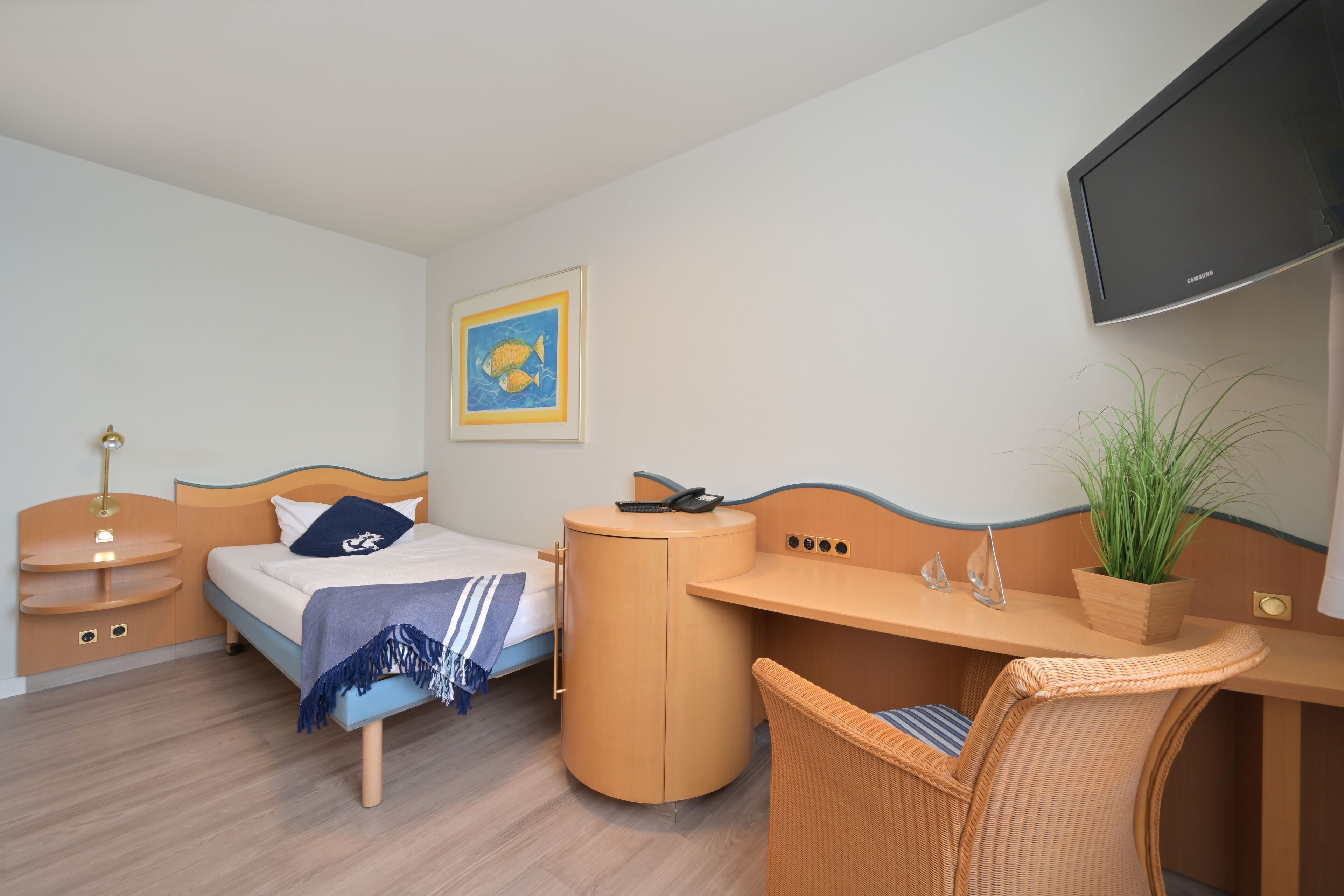 A compact hotel room with a bed, bedside table, desk, chair, TV, and a potted plant.