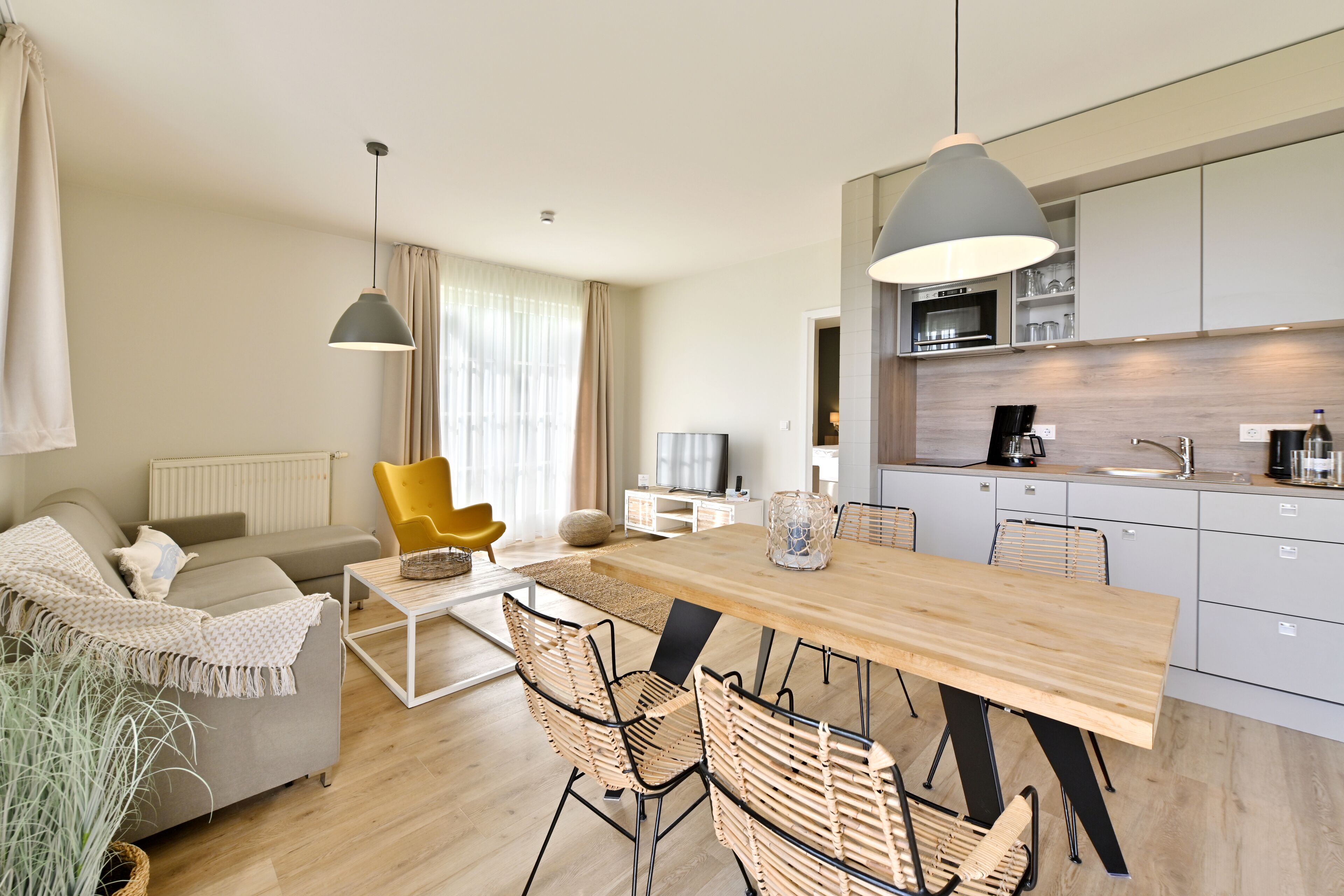 Uma sala de estar moderna com sofá, poltrona amarela, mesa de jantar e cadeiras. Uma cozinha com armários e eletrodomésticos é visível ao fundo.