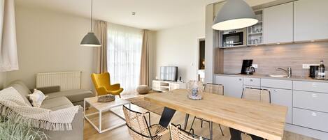 Uma sala de estar moderna com sofá, poltrona amarela, mesa de jantar e cadeiras. Uma cozinha com armários e eletrodomésticos é visível ao fundo.