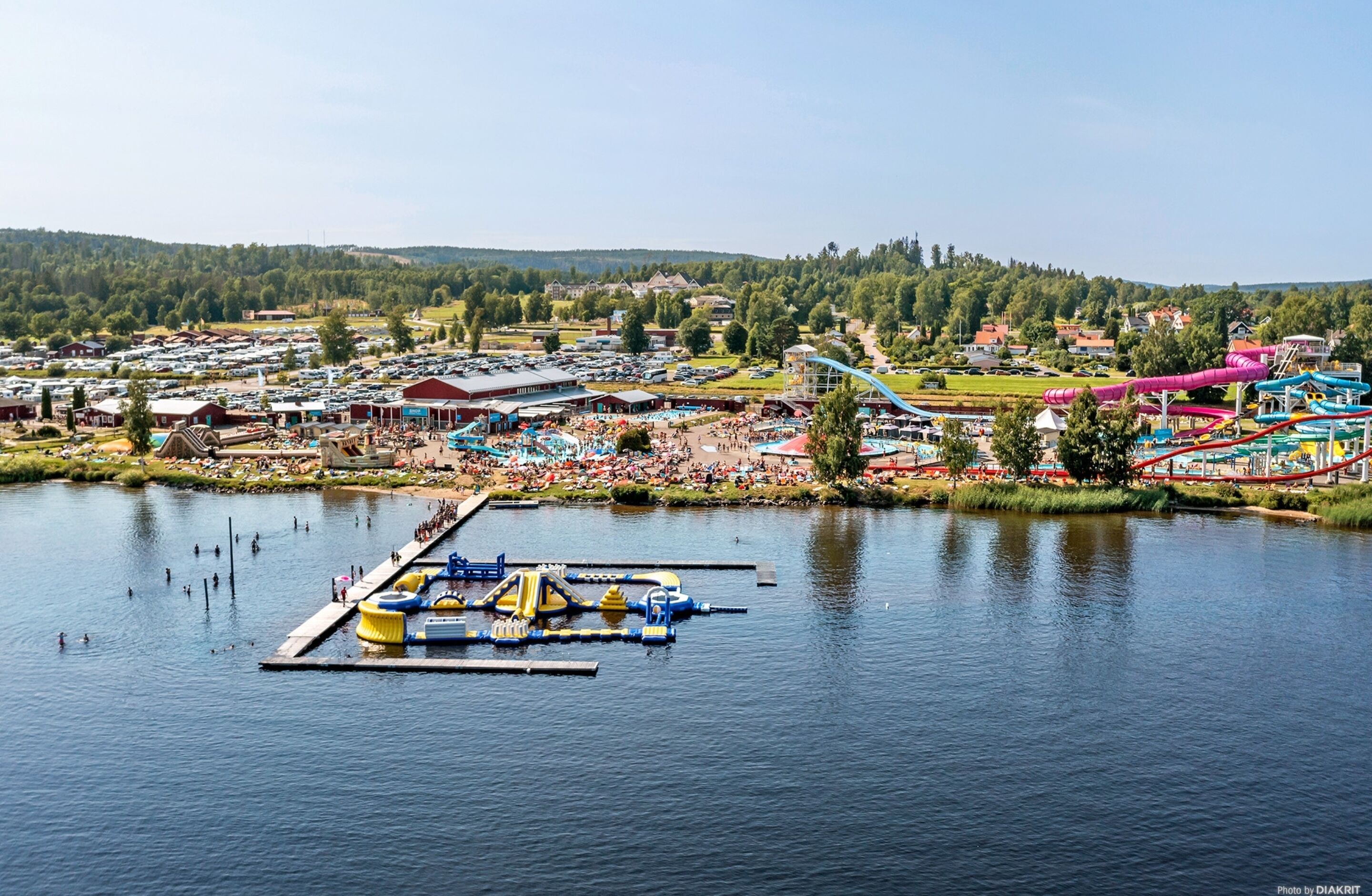 Huvipuisto järven rannalla, jossa on vesilaitteita, laituri, jossa on käytössä puhallettavia veneitä, ja paljon ihmisiä.