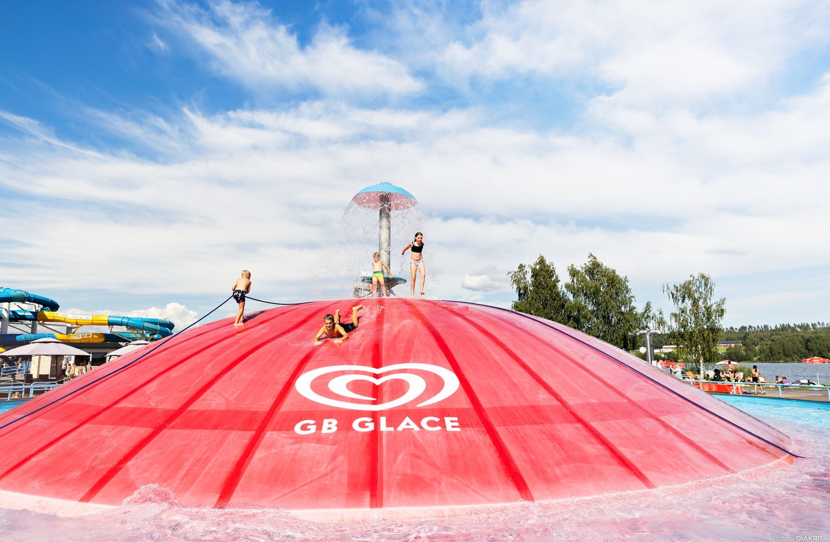 A water slide with the GB GLACE logo, surrounded by a pool area with umbrellas and people.