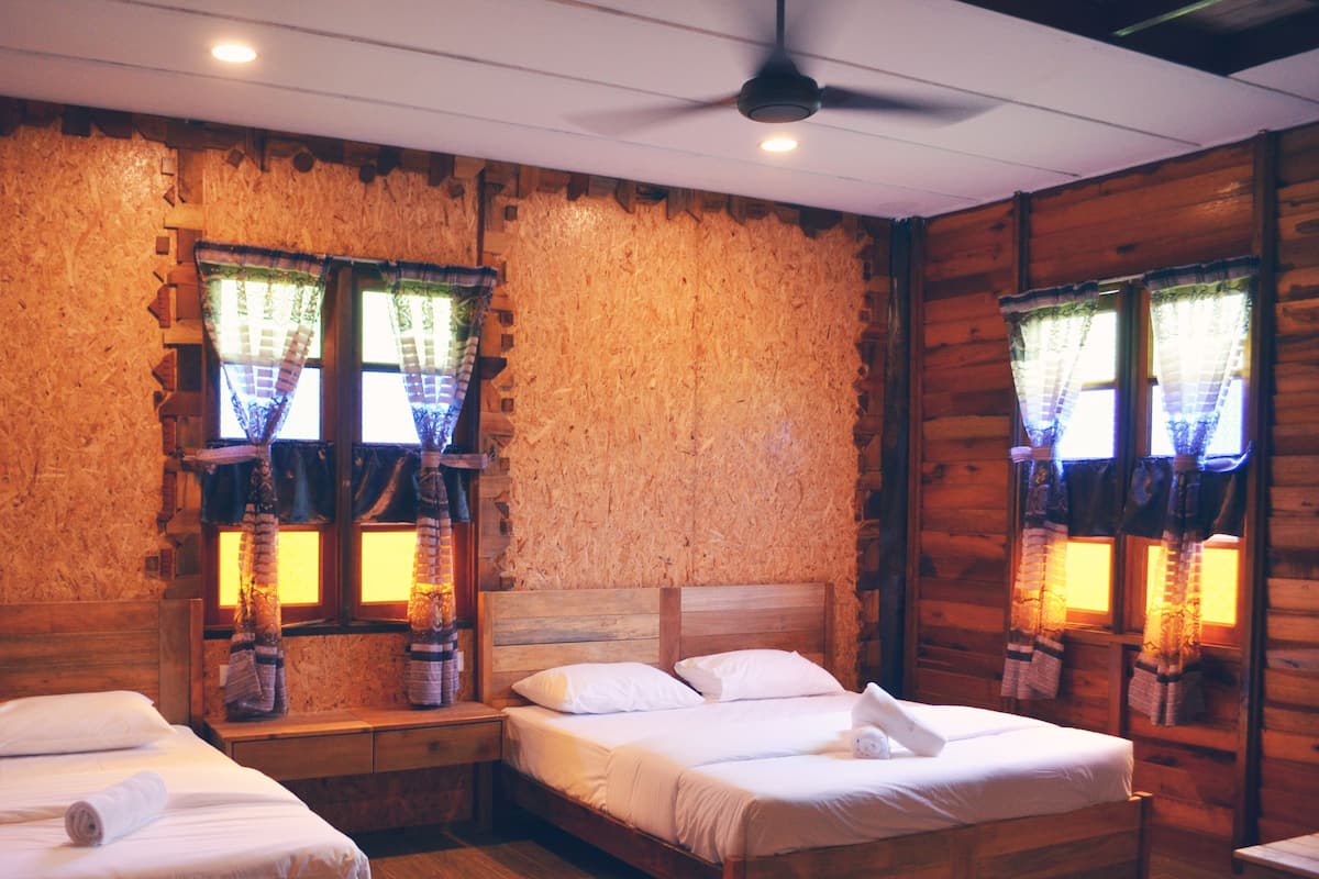 Una habitación tipo cabaña de madera con dos camas individuales, un ventilador de techo y ventanas amplias con cortinas.