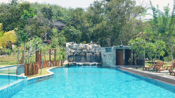 A swimming pool with a waterfall feature, surrounded by lush greenery and wooden deck chairs. - The Acacia Retreat (Bentong)