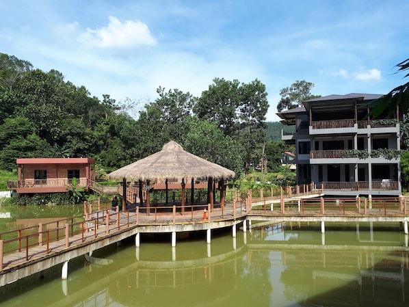 A thatched-roof pavilion, a wooden bridge over a pond, and a multi-story building with balconies.