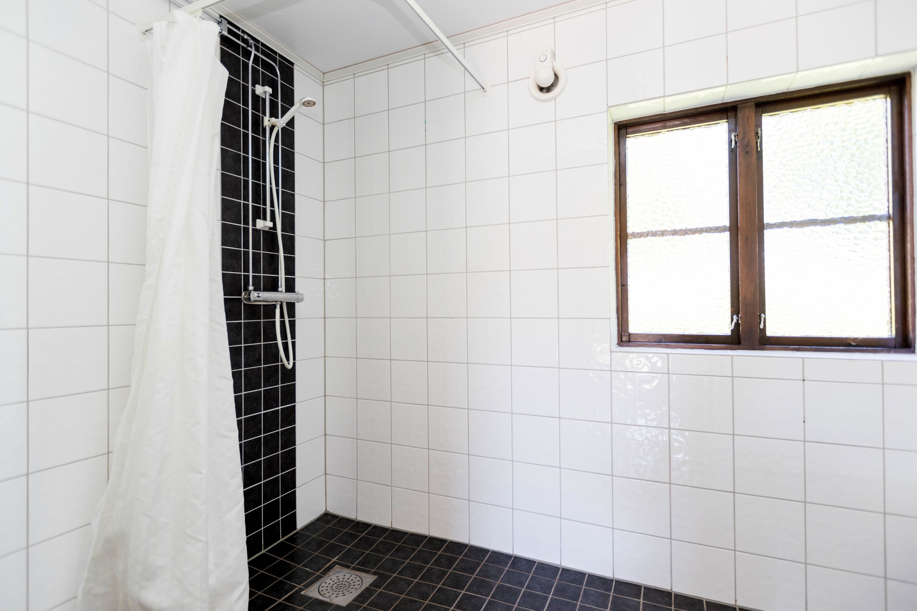 Ein Badezimmer mit weißen Fliesen an den Wänden, einem schwarzen Duschbereich mit Fliesen, einem weißen Duschvorhang und einem Fenster mit Holzrahmen.