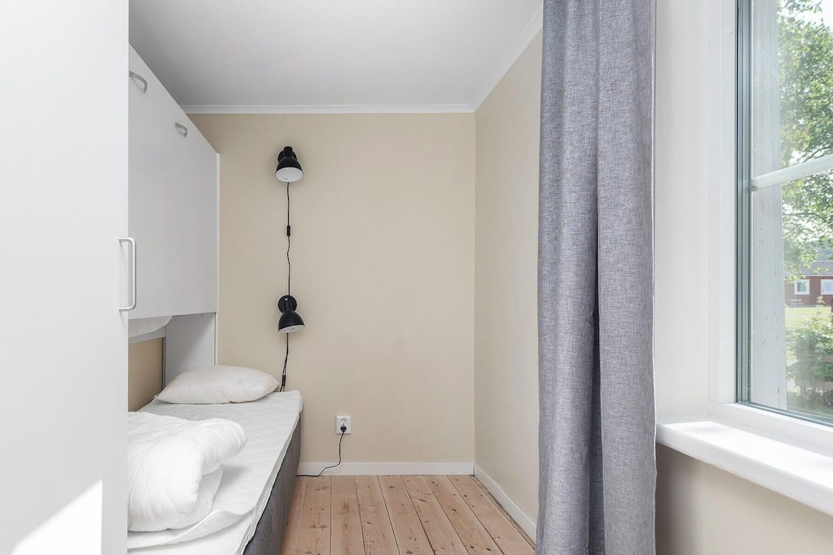 A small bedroom with a bed, a nightstand, a lamp, and a window with curtains.