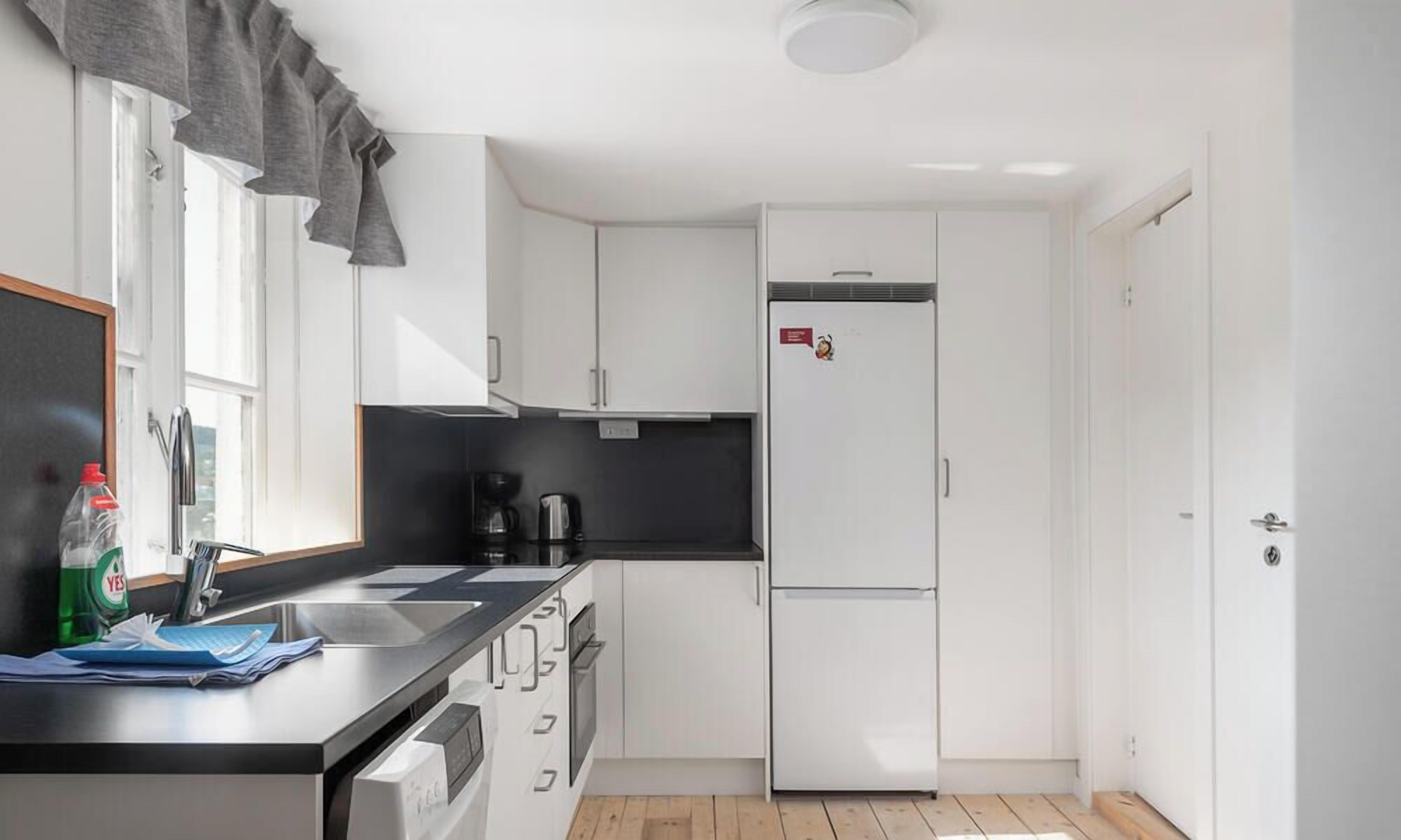 Cocina compacta con armarios blancos, encimera negra, refrigerador y ventana con cortinas.
