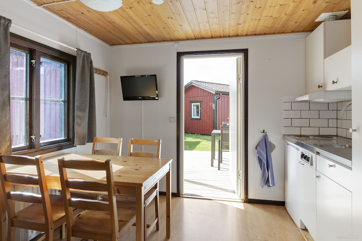 A kitchen with a wooden ceiling, a dining area with a table and chairs, a TV mounted on the wall, and a view of a red building outside.