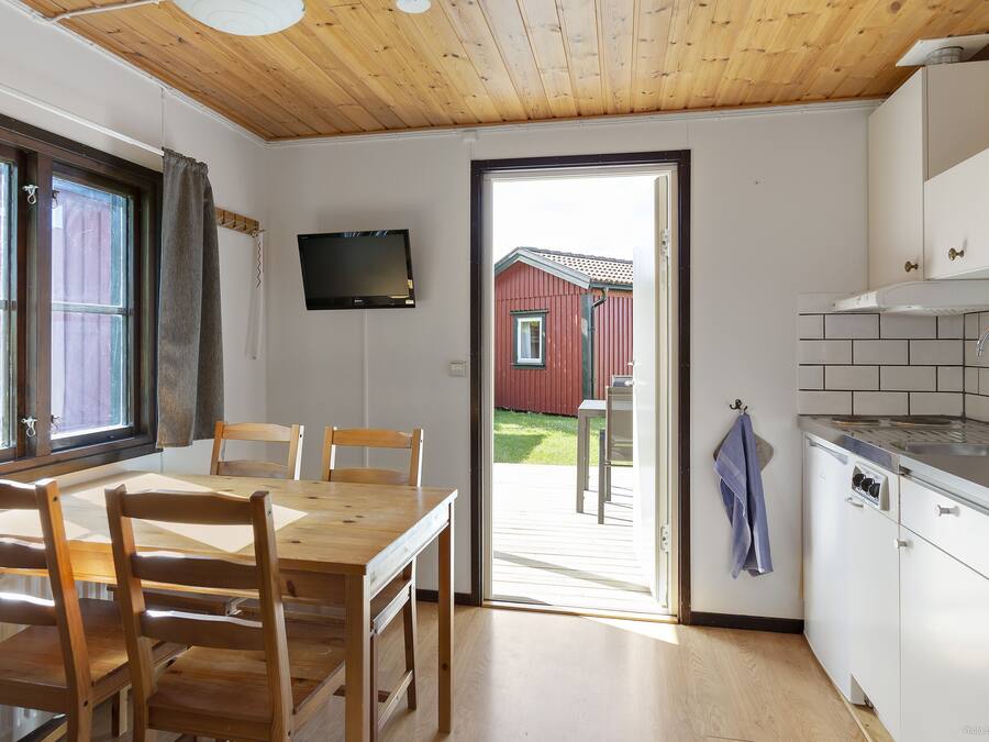 Een keuken met een houten plafond, een eethoek met een tafel en stoelen, een aan de muur bevestigde televisie, en uitzicht op een rood gebouw.