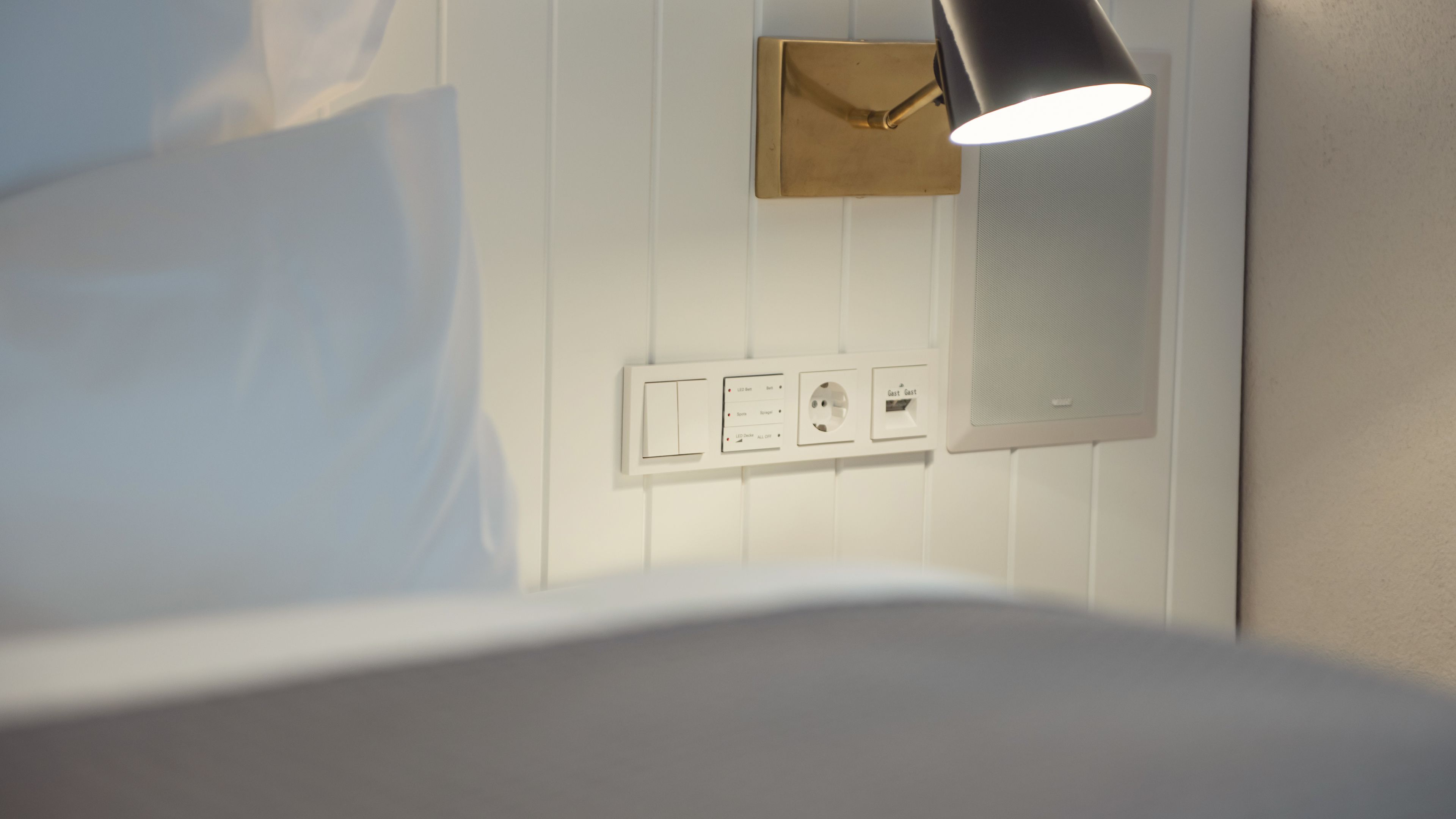 Quarto de hotel moderno com cama, luminária na parede e tomada com interruptor.