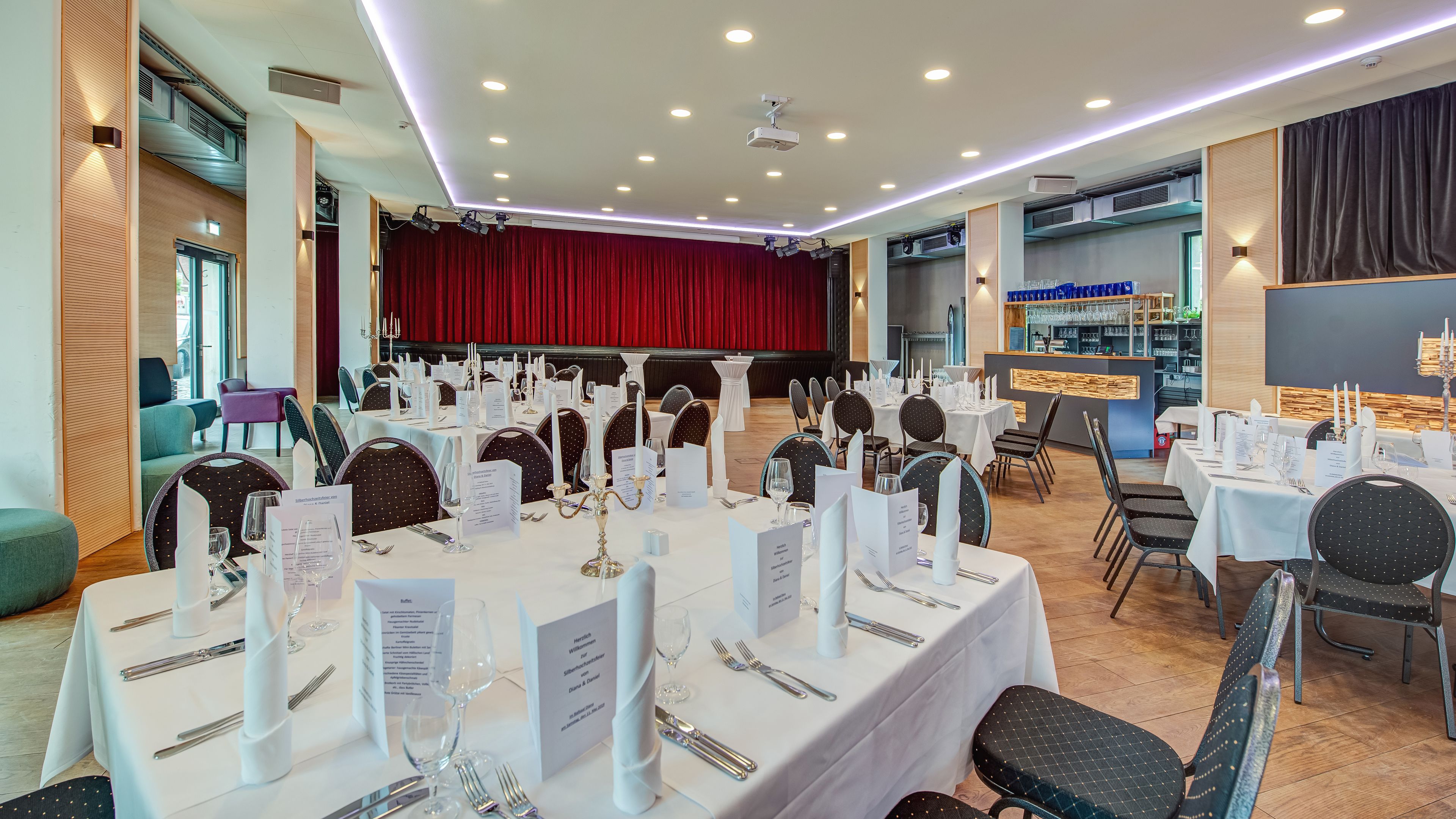 A banquet hall with round tables set for dining, a stage with red curtains, and a bar area with shelves of glasses.