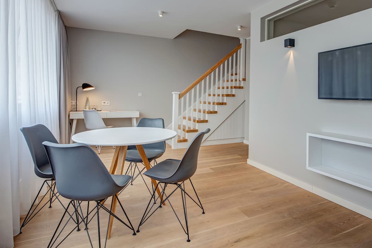 A modern living room with a round dining table, grey chairs, a staircase, a mounted TV, and a white console unit.