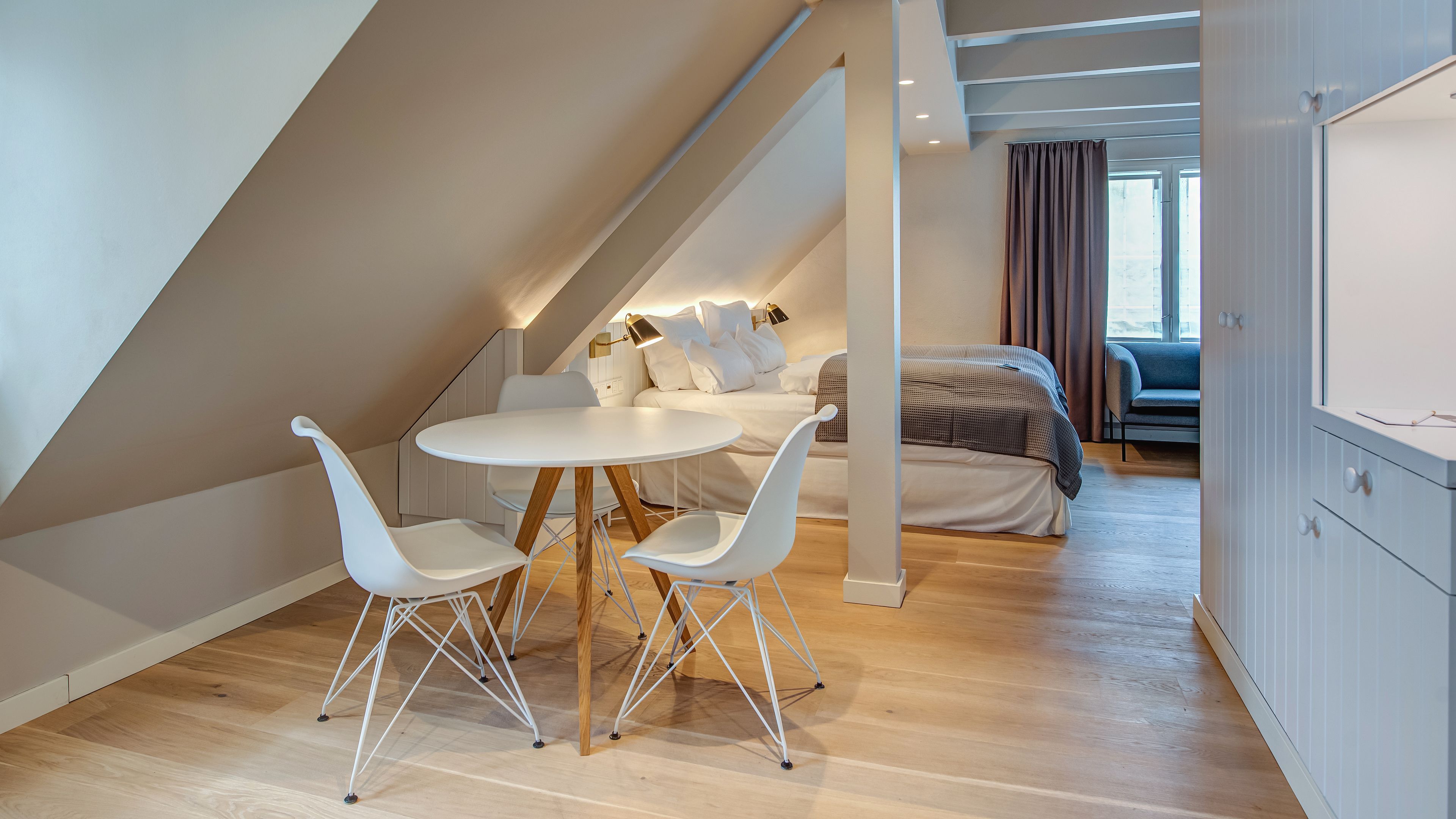 Una moderna camera da letto mansardata con soffitto spiovente, un tavolo rotondo con sedie bianche, un letto con copripiumino bianco e grigio a quadri, e vista all'esterno tramite una finestra con tende.
