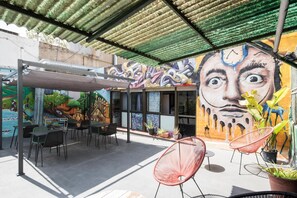 An outdoor seating area with tables and chairs, a mural with abstract art, and a covered patio with a green roof. - No Limit Hostel Graffiti (Barcelona)