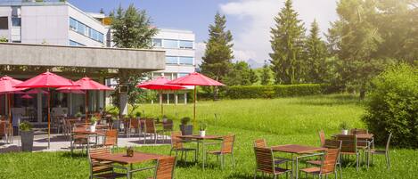 An outdoor cafe with tables and chairs, red umbrellas, and a building in the background.