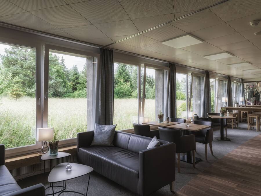 A modern dining area with a long table, chairs, and a sofa. Large windows provide a view of a grassy area.