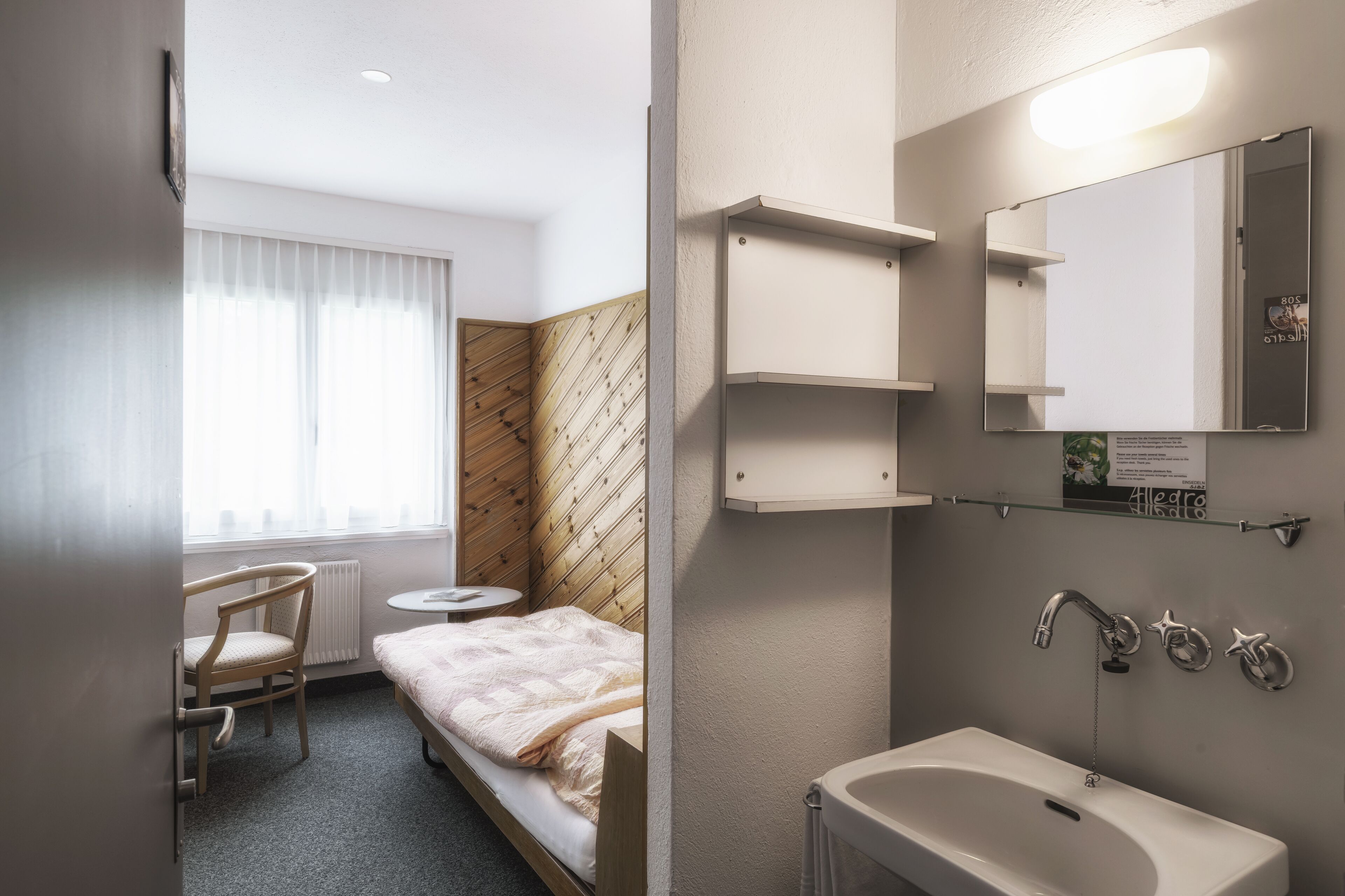 Une chambre d’hôtel comprenant un lit, une chaise, une petite table, une fenêtre avec des rideaux, un mur d’accent en bois, une salle de bain avec un lavabo, un miroir et des étagères.