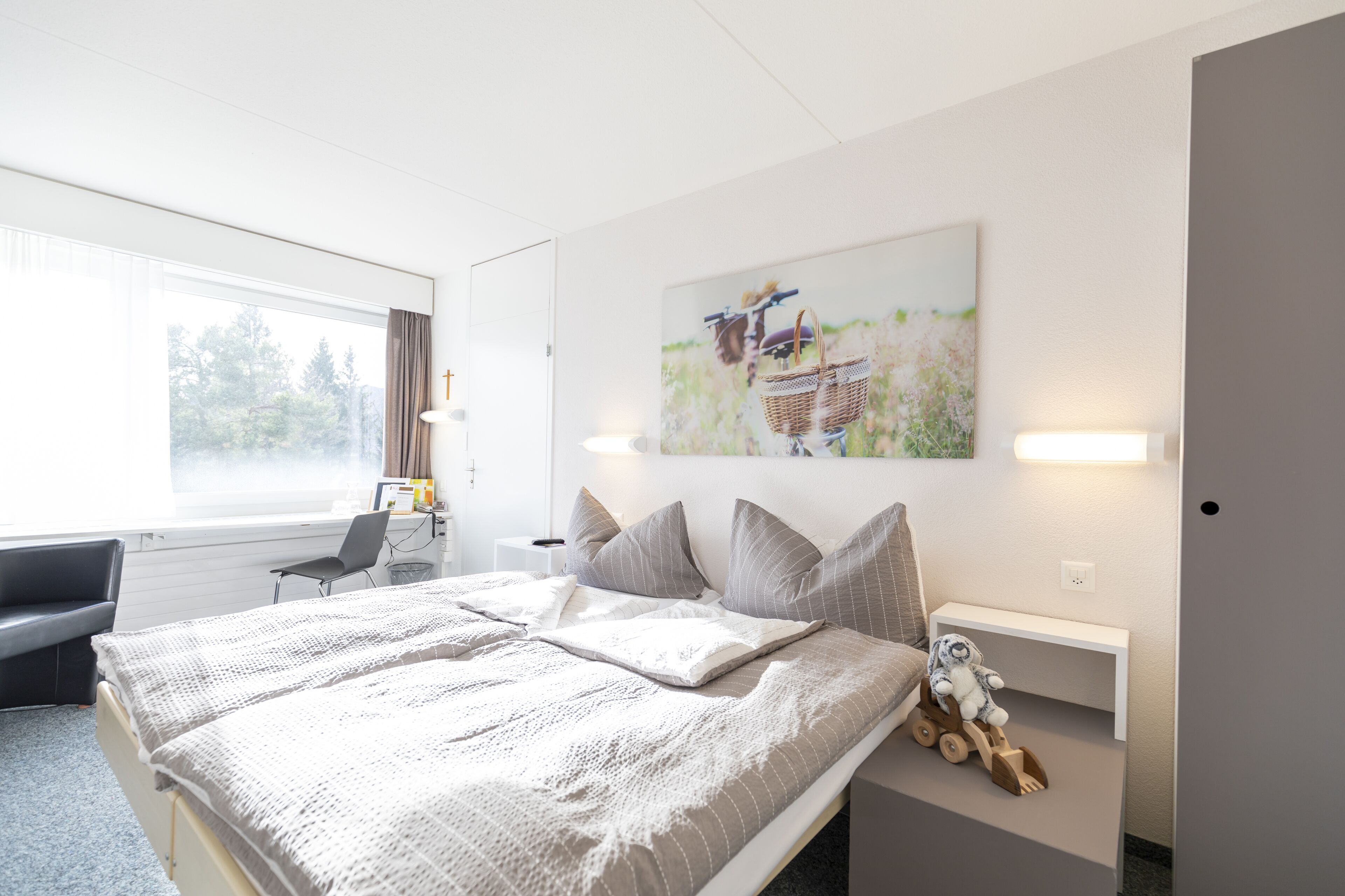 Moderni hotellihuone, jossa on suuri sänky, työpöytä ja tuoli. Huoneessa on ikkuna, josta avautuu näkymä puita kohti, ja työpöydällä on lamppu.