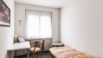 Une petite pièce bien éclairée, avec un lit simple, un bureau avec une chaise, une petite table et une fenêtre avec des rideaux.