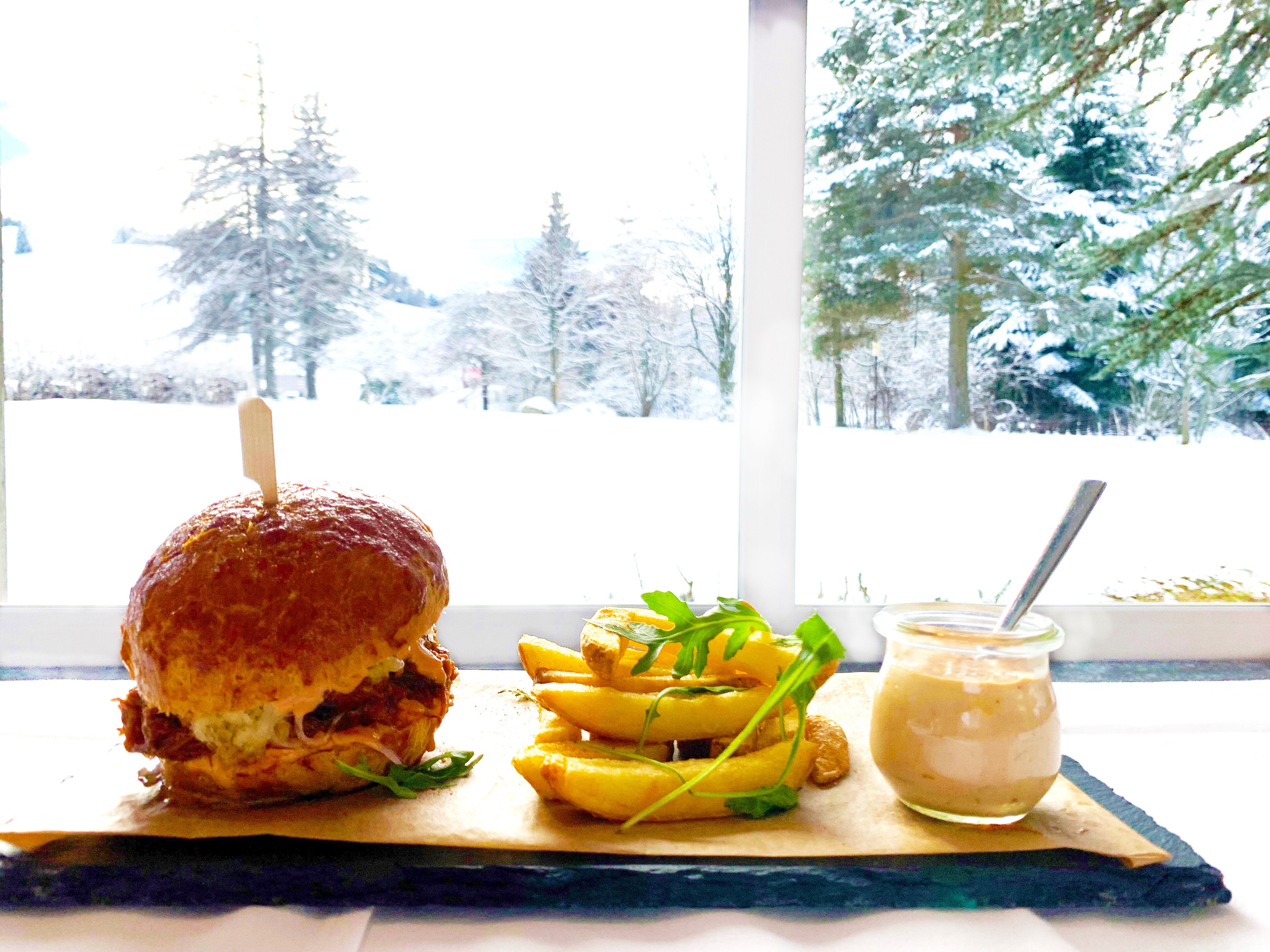 Un sándwich, papas fritas y un recipiente de salsa sobre una tabla de madera junto a una ventana con vistas nevadas.