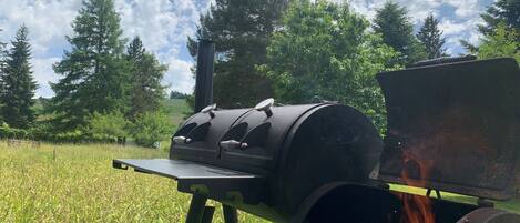 Una parrilla portátil con una plancha superior y rejillas laterales, ubicada en un campo cubierto de césped con árboles al fondo.