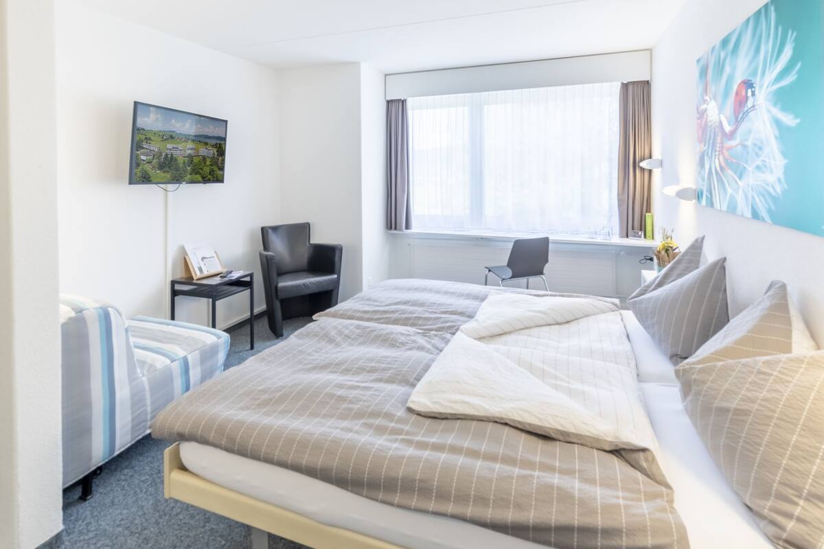 A hotel room with a large bed, a desk, a chair, a TV, and a window with curtains.