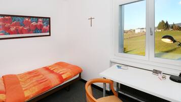 Een kamer met een bed, een bureau, een stoel, een raam met uitzicht op een grasveld, en een kruis aan de muur.