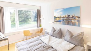 Un dormitorio con cama, escritorio, silla, ventana con cortinas y un cuadro con una escena portuaria.