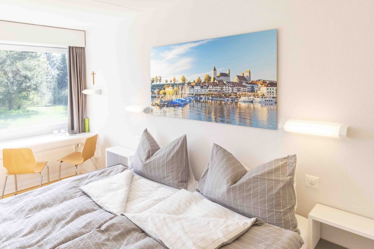 A bedroom with a bed, a desk, a chair, a window with curtains, and a painting of a harbor scene.