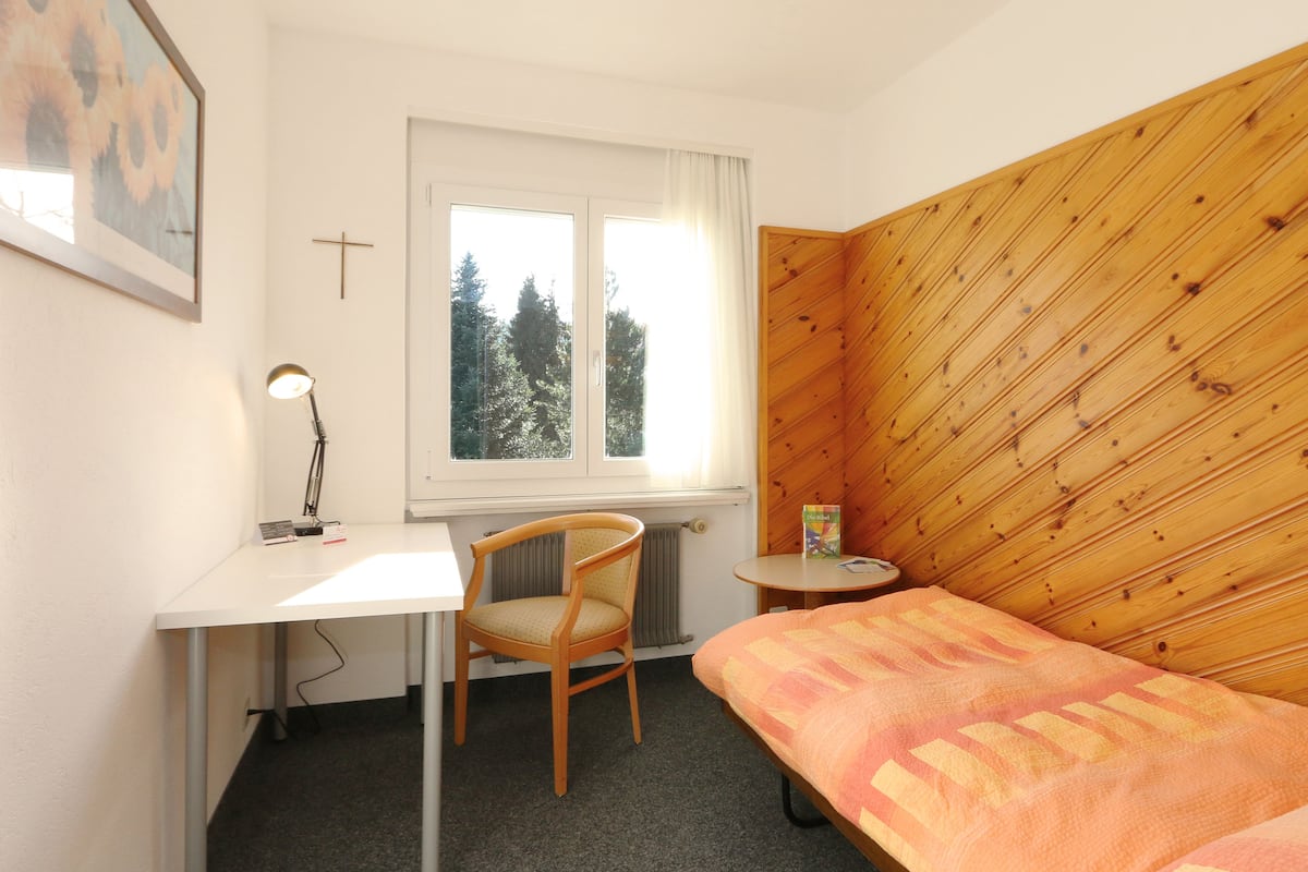 A bedroom with a wooden headboard, a desk with a chair, a small bedside table, and a window with a view of trees.