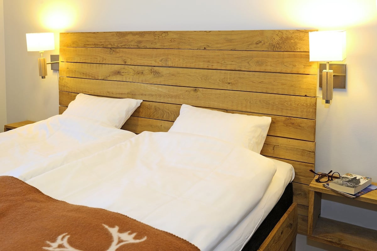 A bed with a wooden headboard, two bedside lamps, and a nightstand with glasses and a book.