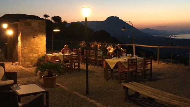 An outdoor dining area with tables and chairs, a stone wall, and a view of the mountains and water in the background. - Agriturismo Case Tabarani (Collesano)