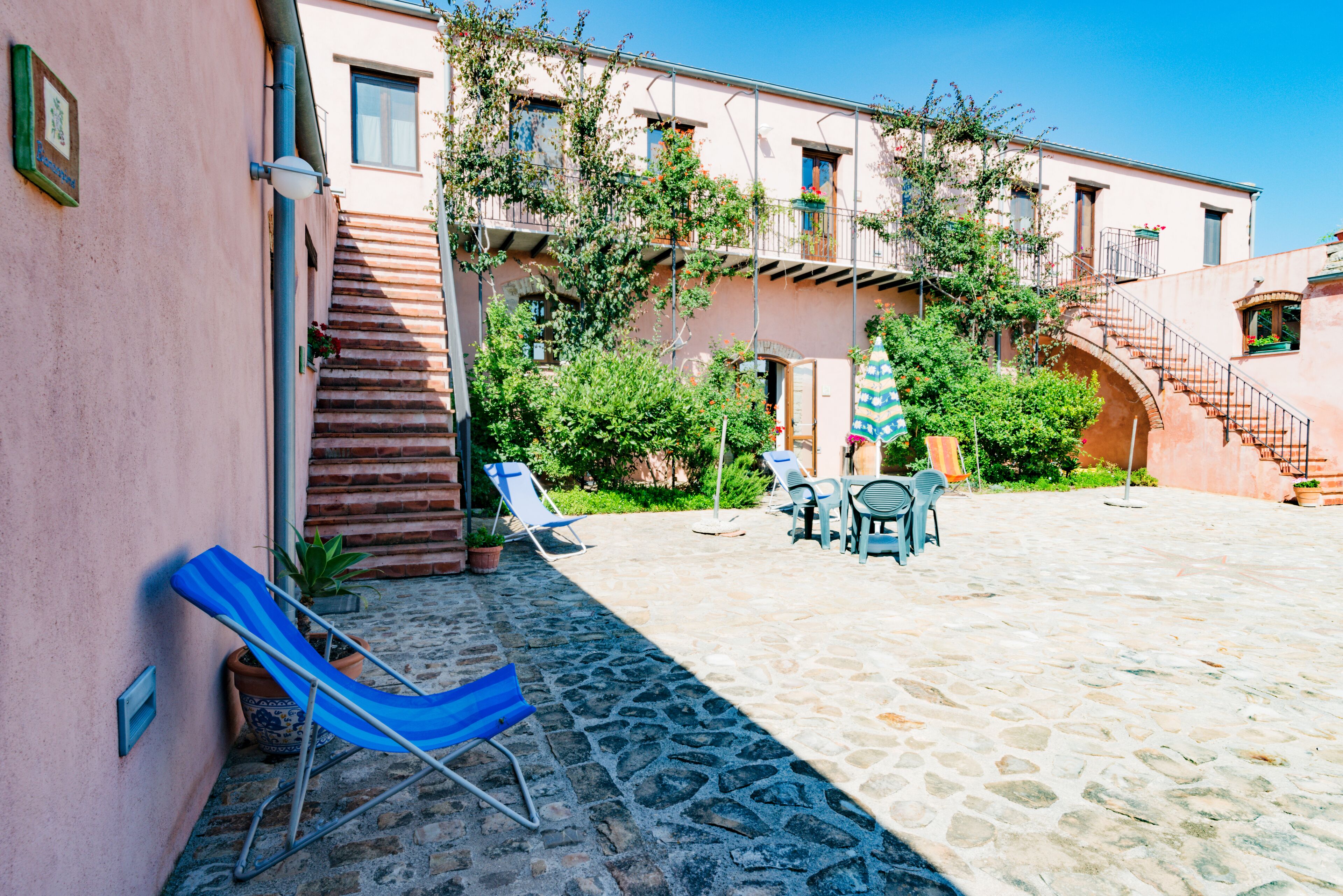 A courtyard with stone flooring, a blue lounge chair, a table with chairs, and a staircase leading to an upper floor.
