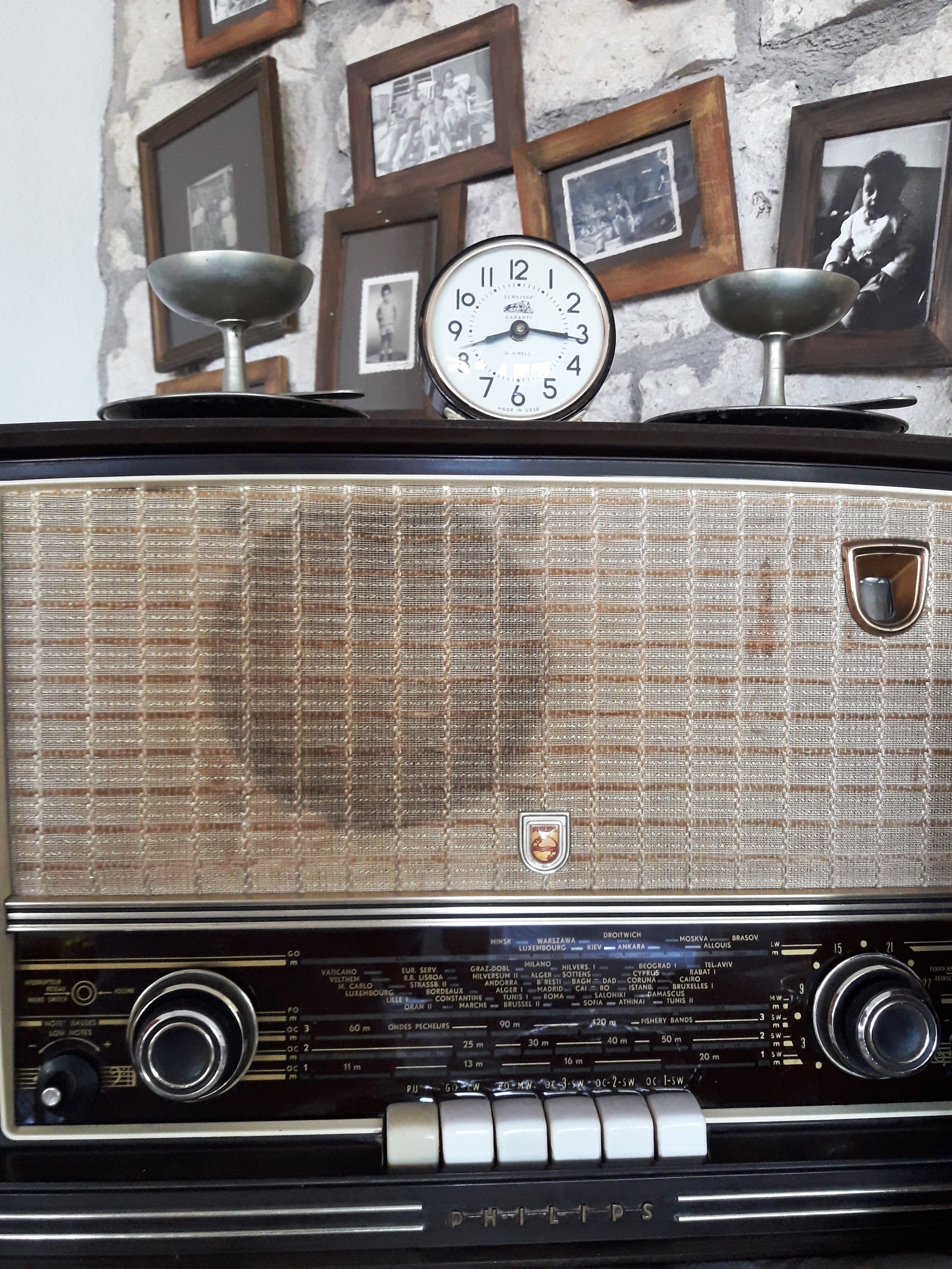Una radio antigua con un reloj encima, frente a una pared texturizada con cuadros enmarcados.