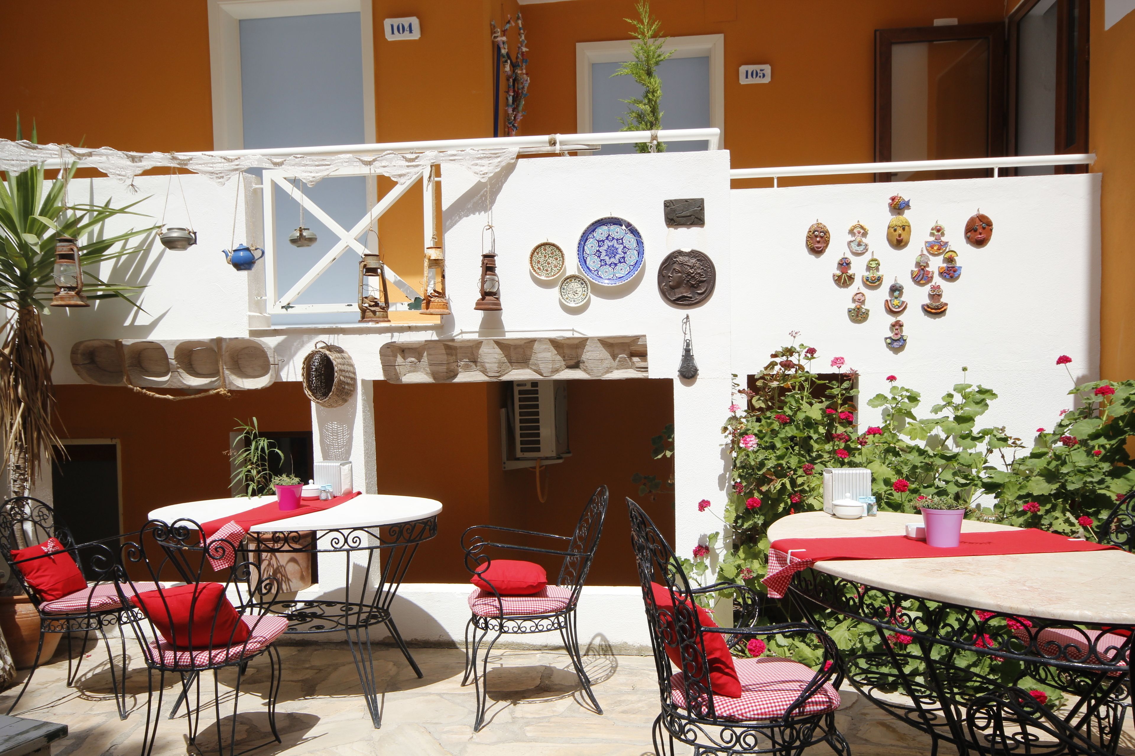 Un patio exterior con mesas y sillas, rodeado de plantas en macetas y platos decorativos.
