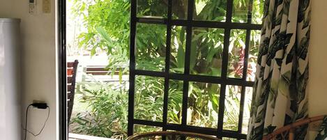 A room with a bed, an exit sign, a window with a view of greenery, and a chair by the window.