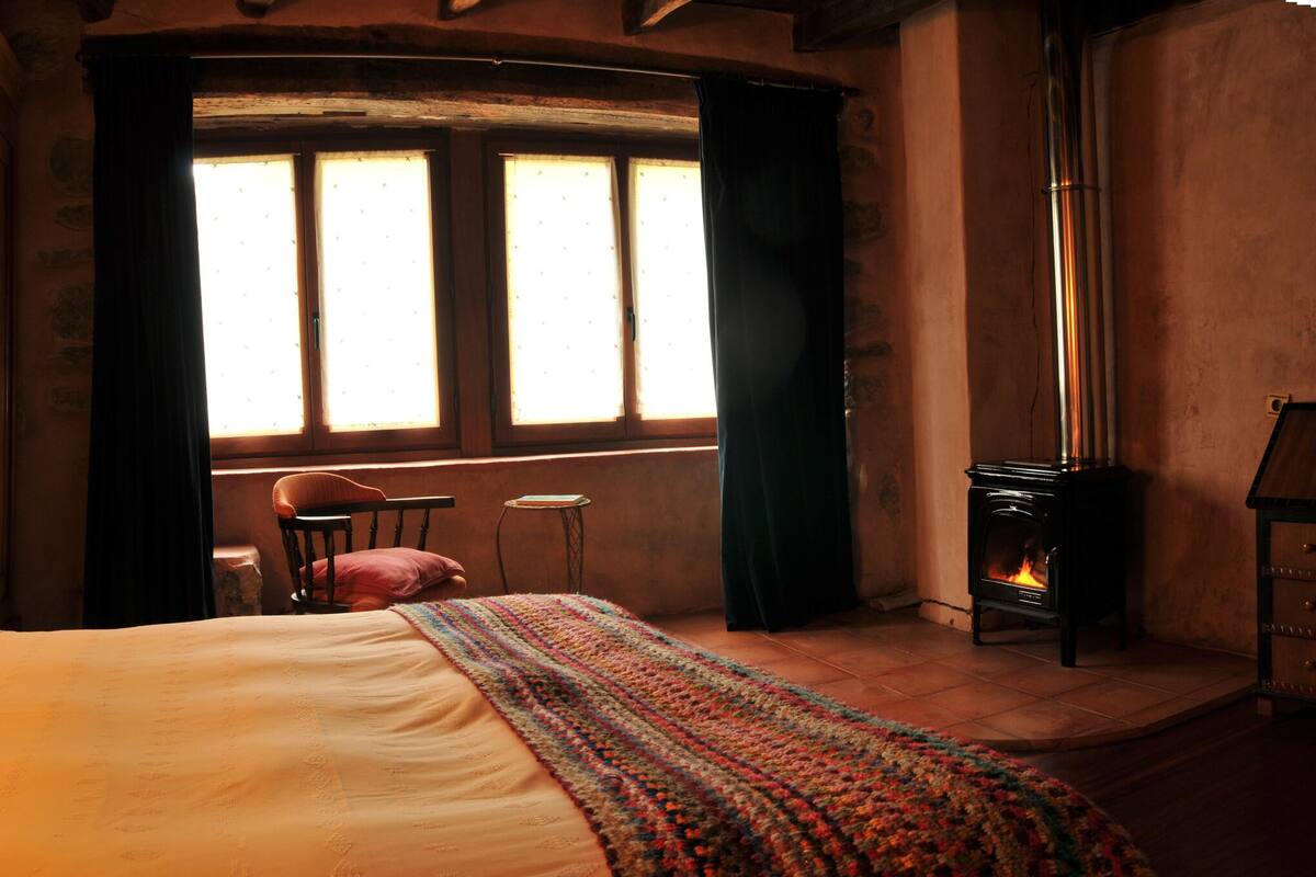 Une chambre confortable avec un lit, deux lampes fixées au mur, une fenêtre avec des rideaux, une petite table et une cheminée.