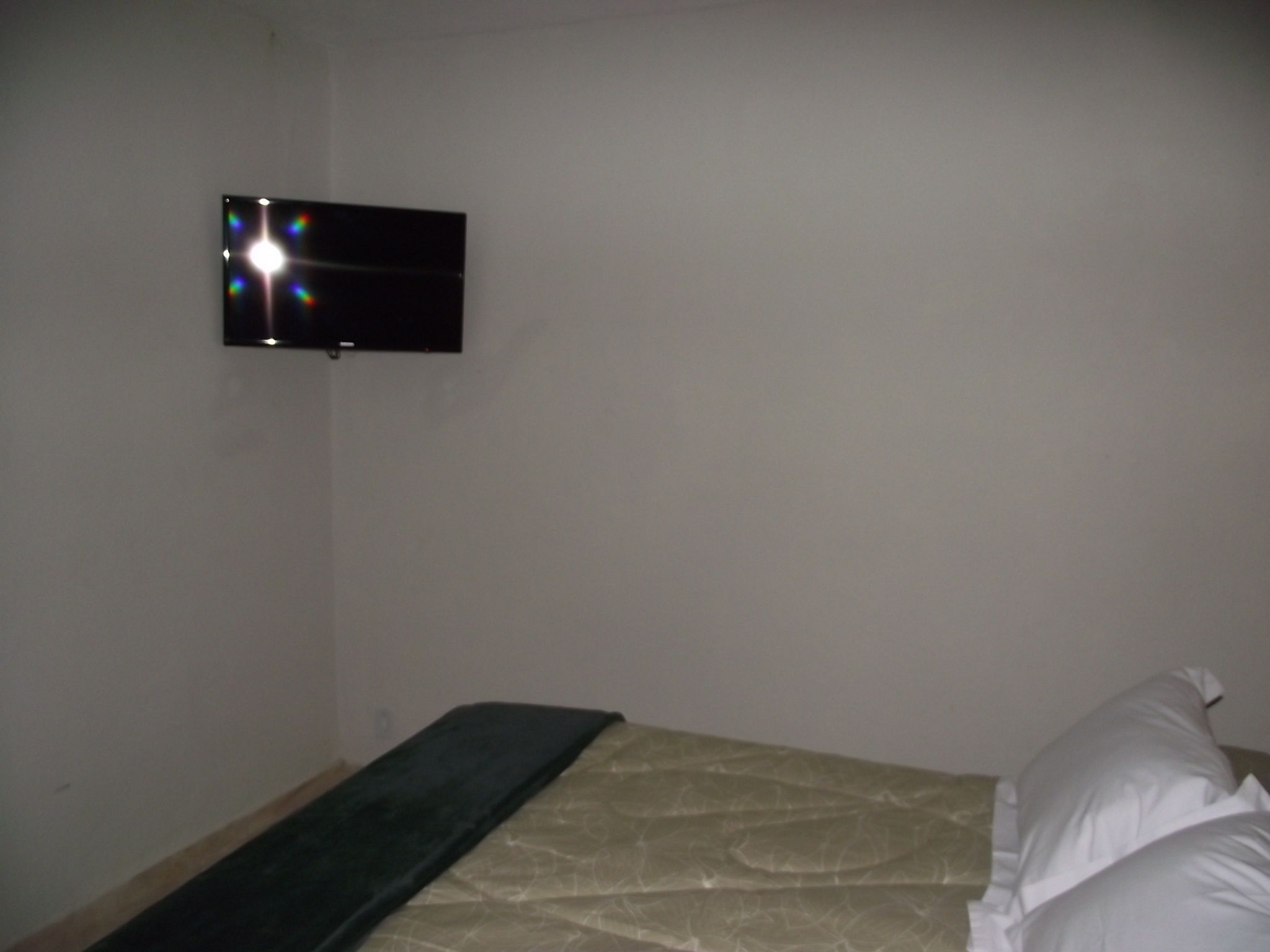 Een kleine, zwak verlichte kamer met een enkel bed, een aan de muur bevestigde televisie en een effen witte muur.
