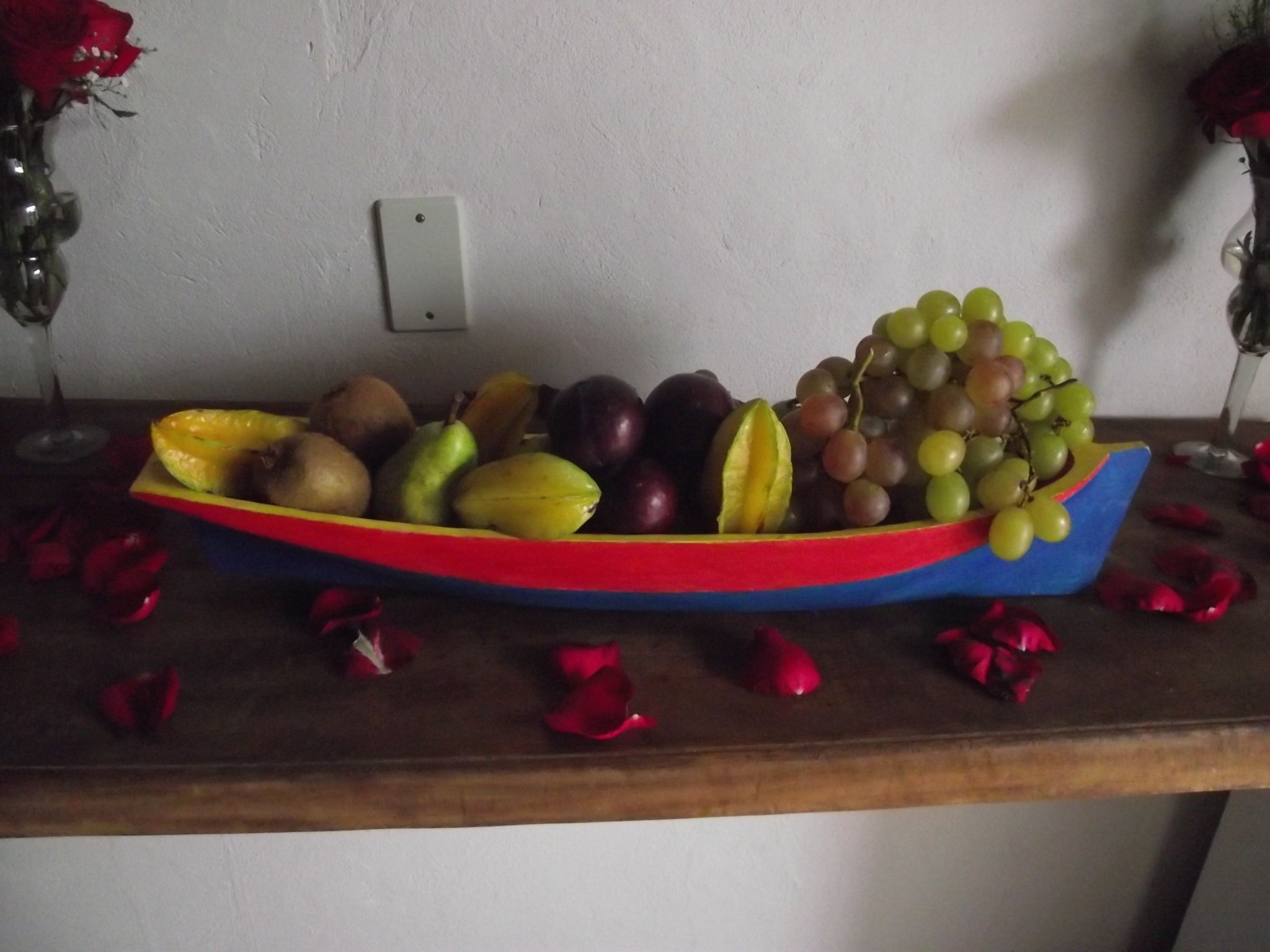 A fruit bowl on a wooden table with rose petals scattered around.
