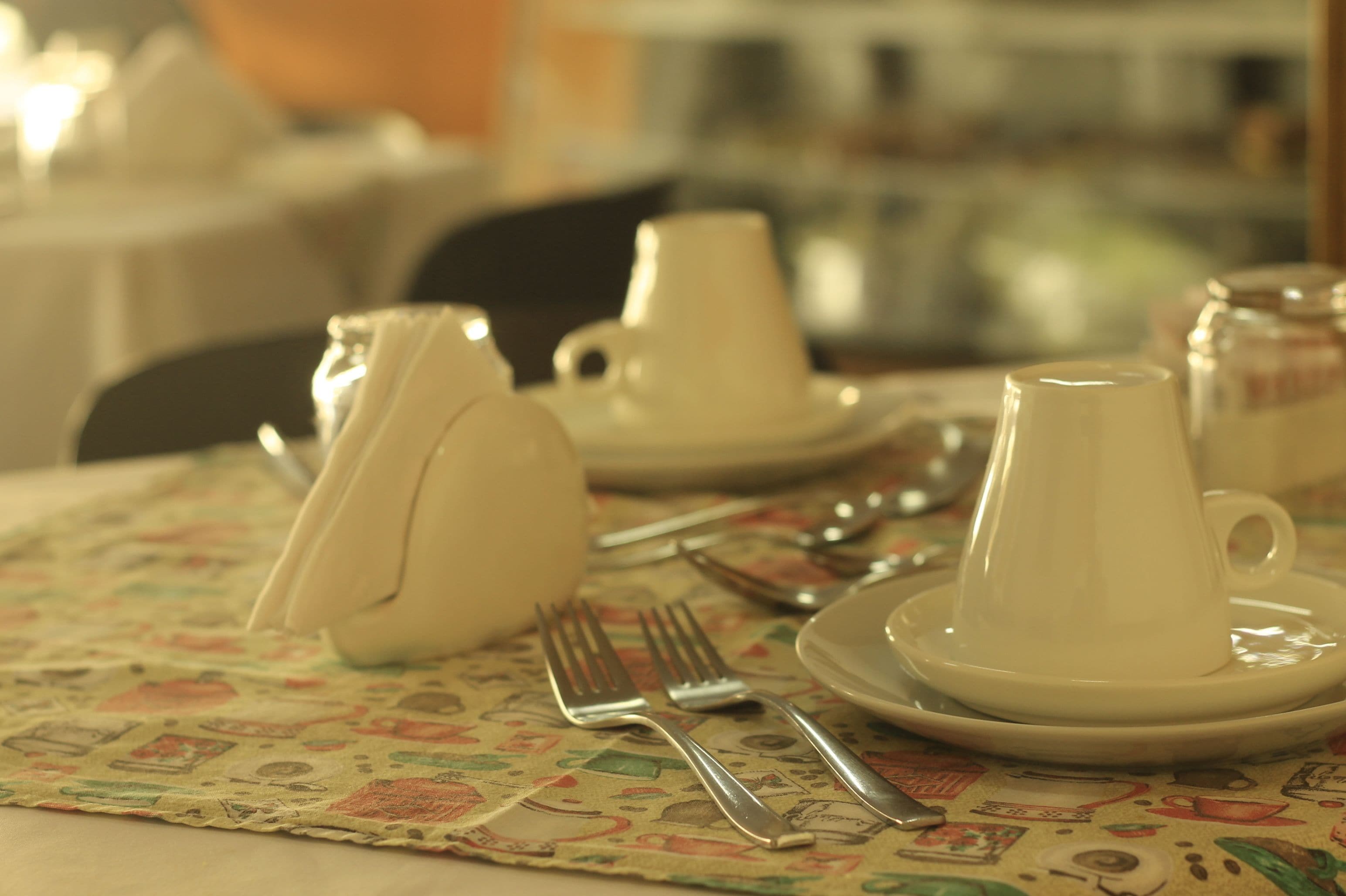 Una mesa servida con tazas, platillos y cubiertos sobre un mantel estampado.