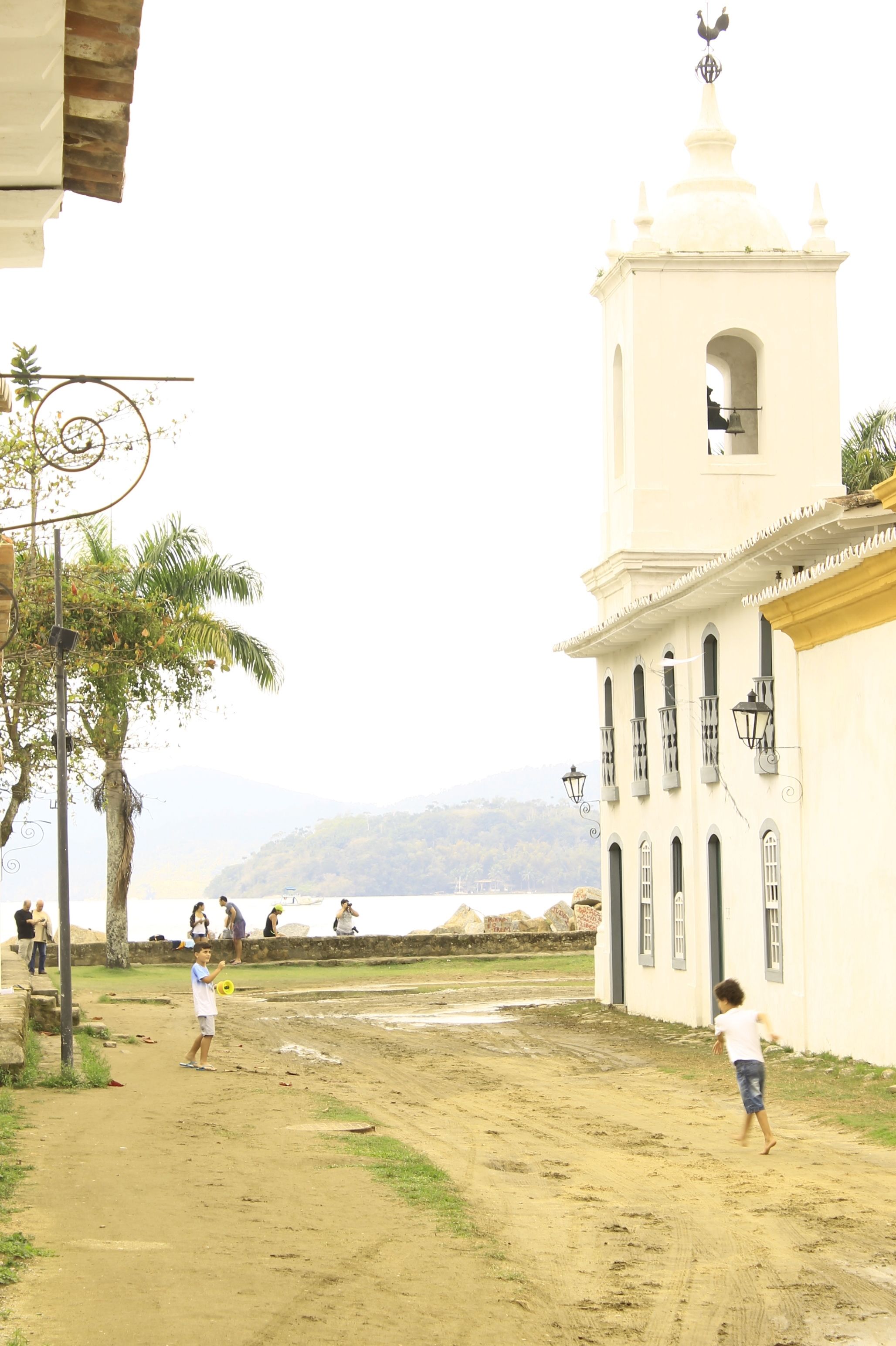 A sandy path leading to a white building with a bell tower, palm trees, and a view of the sea.
