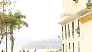 A sandy path leading to a white building with a bell tower, palm trees, and a view of the sea.