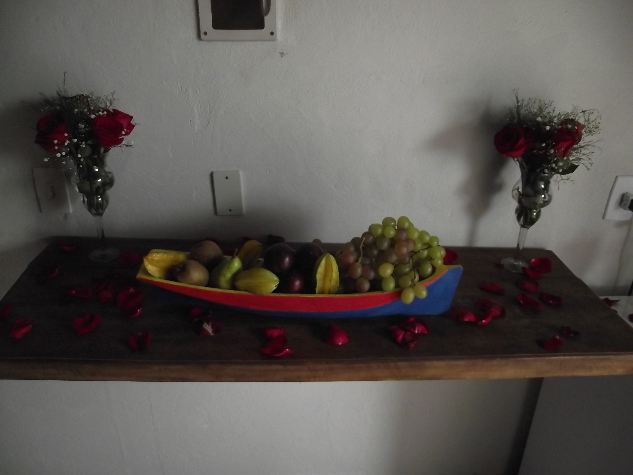 Una mesa con un tazón lleno de fruta colorida y un florero con rosas rojas a cada lado.