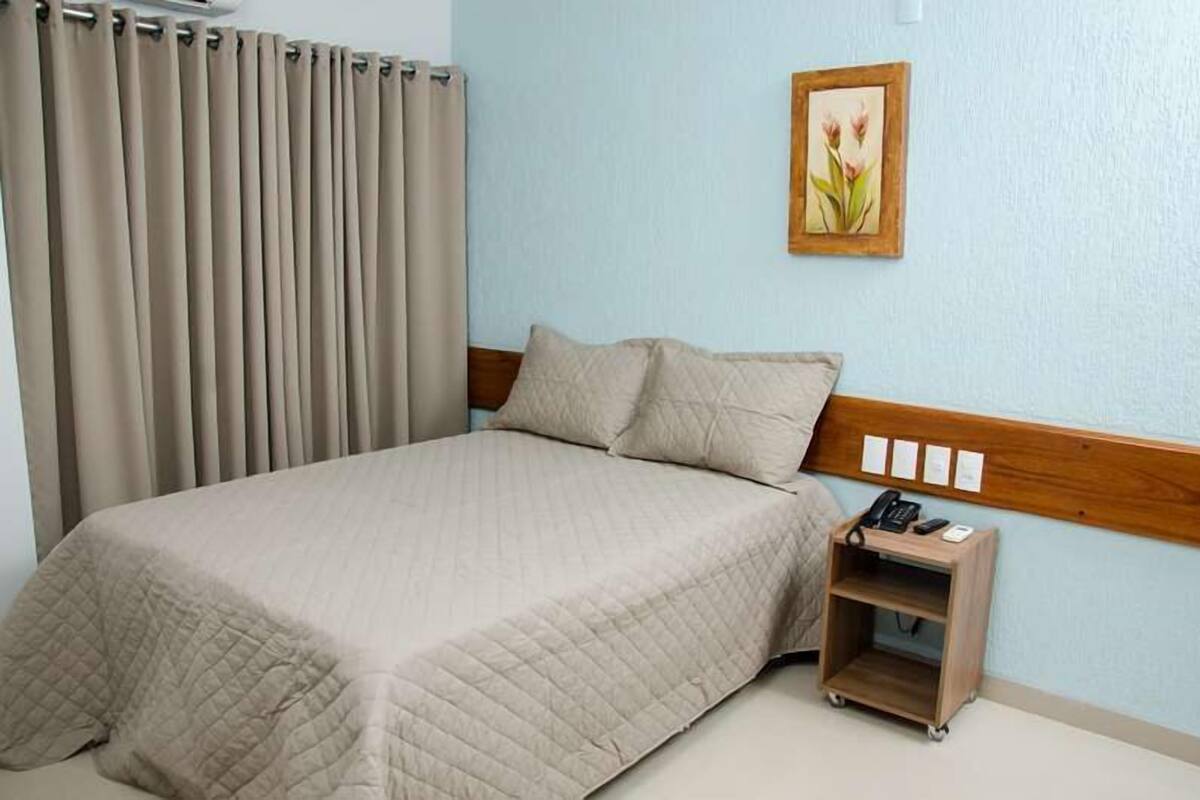 A neatly made bed with a beige quilt, a wooden headboard, a bedside table with a phone and a small shelf, and a light blue wall with a floral painting.