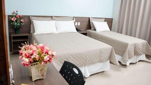 A hotel room with two beds, a desk, and a chair. There are floral arrangements on the desk and a nightstand with a phone. - Serras Hotel (Rondonopolis)