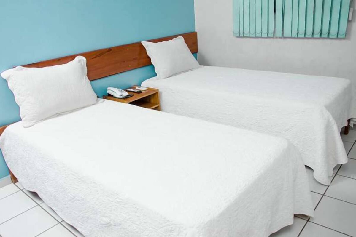 A room with two single beds, each with a wooden headboard and white bedding. There is a small wooden nightstand with a telephone and a lamp between the beds.