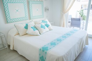 Une chambre avec un grand lit, du linge de lit blanc et des coussins décoratifs de couleur turquoise. On y trouve deux appliques murales et une fenêtre avec des rideaux.