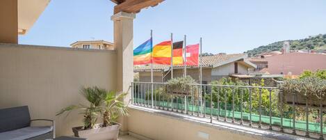 A balcony with a sofa, a small table, and a potted plant. There are two flags on the railing.