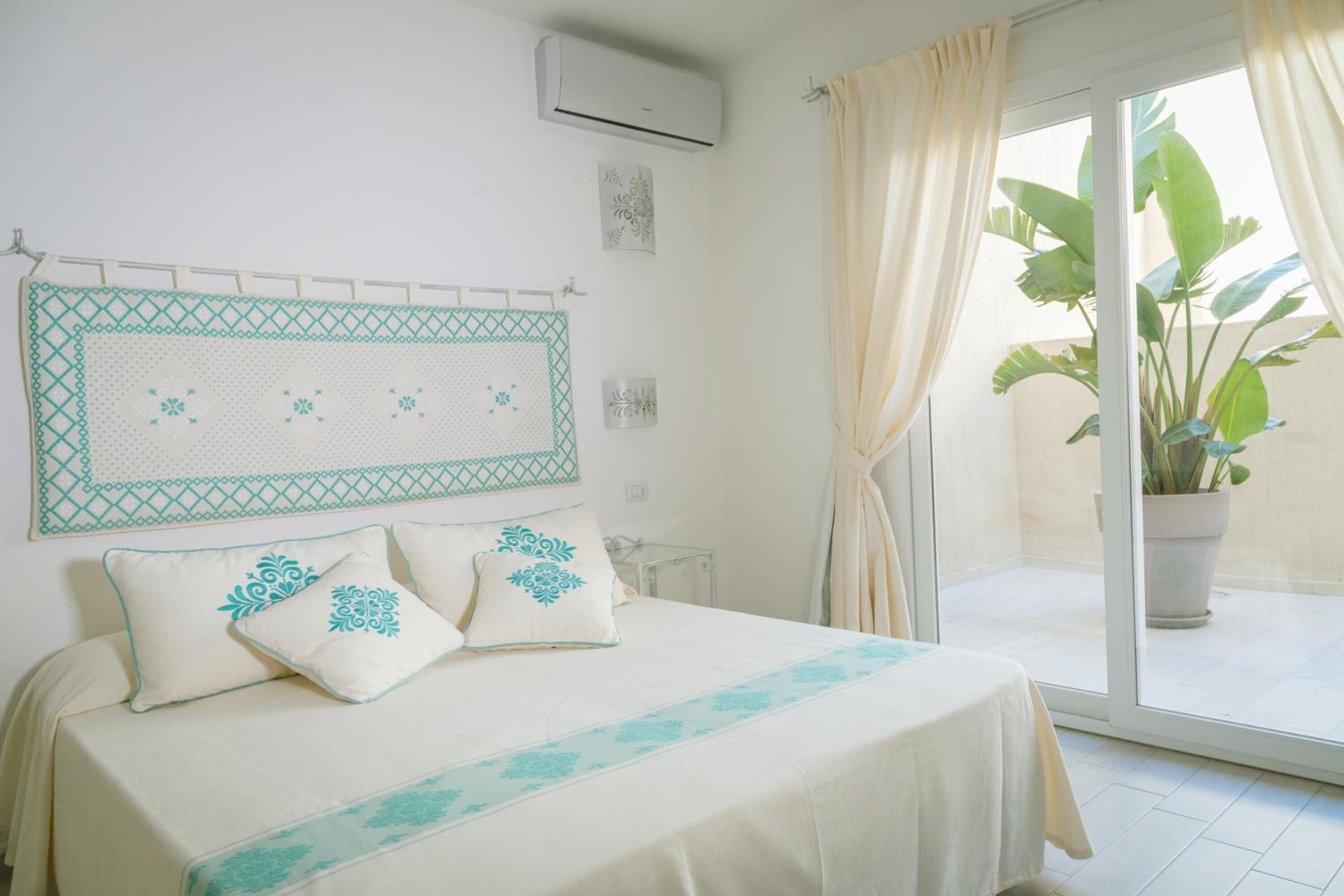 A bedroom with a bed, pillows, a wall-mounted air conditioner, and a view of a balcony with a plant.