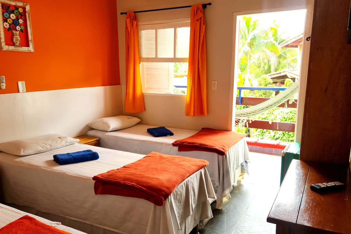 A room with two beds, each with a white mattress and orange towels. There is a window with orange curtains, a hammock outside, and a remote control on a wooden table.